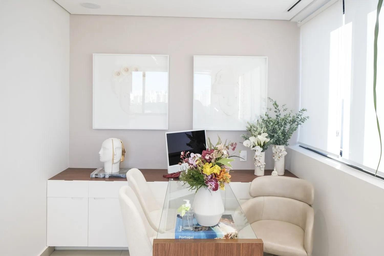 Sala do consultório com mesa de vidro, cadeiras, aparador branco e arranjos de flores
