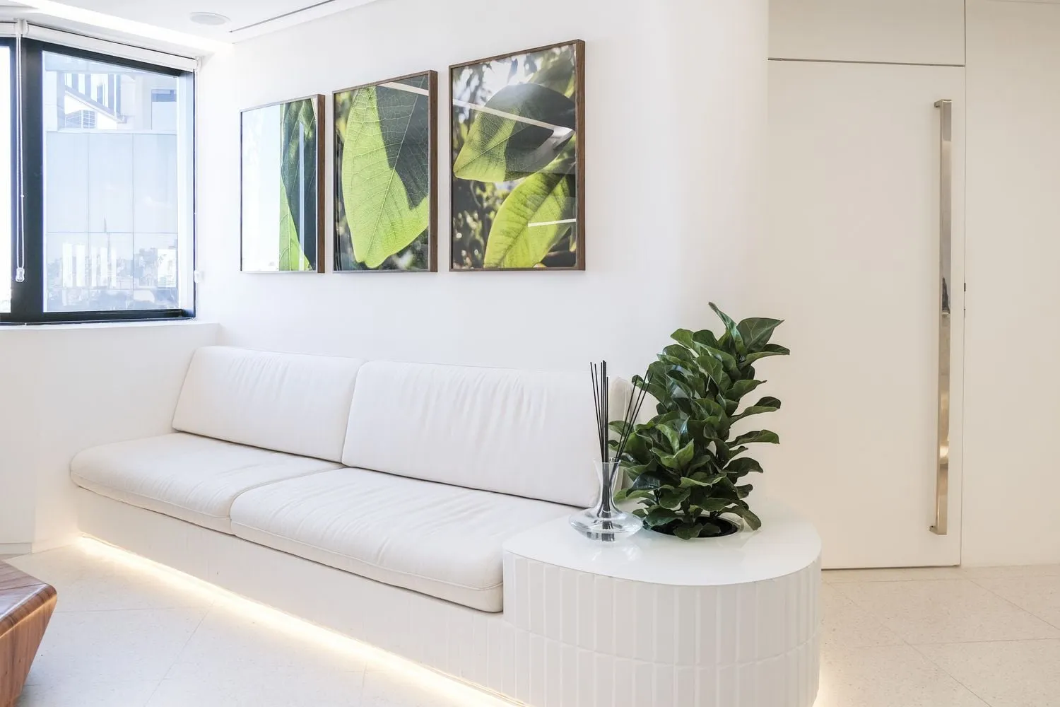 Sala de espera com sofá branco, quadros de folhagens e iluminação indireta